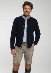 Marineblauwe gebreide cardigan met ronde hals en metalen knopen, gecombineerd met een lichtblauw shirt en beige shorts met blauwe borduursels.