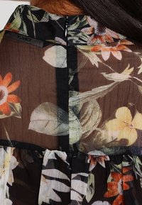 Blouse transparente à motifs floraux avec un fond noir orné de fleurs colorées, un col montant et une fermeture éclair au dos. Tissu texturé.
