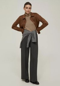 Giacca in finta pelle marrone, dolcevita a righe, pantaloni grigi a gamba larga e un pullover grigio legato in vita. Scarpe nere completano l'outfit.