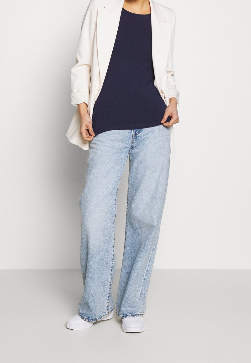Personne portant un jean ample bleu clair, des baskets blanches, un haut bleu marine et un blazer blanc, debout devant un fond clair uni.