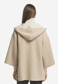 Beige huva poncho med en texturerad, ljusgrå krage. Har vida ärmar och en lös passform, tillverkad av mjukt, varmt tyg.