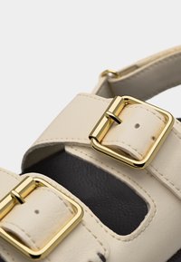 Κοντινή λήψη μπεζ δερμάτινων λουριών σανδαλιού με χρυσές τετράγωνες αγκράφες και μαύρη υφασμάτινη εσωτερική σόλα.