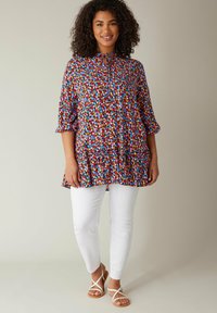 Kleurvolle bloemen tuniek met korte mouwen, rucheshals en strikdetail, gecombineerd met slim-fit witte broeken en platte sandalen.