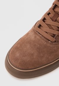 Brown suede sneaker with thick rubber sole and matching brown laces on a plain white background.