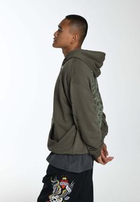 Felpa verde oliva con un grande design grafico sulla schiena, vestibilità comoda, tasca frontale e cappuccio foderato, abbinata a pantaloni neri con ricami colorati.