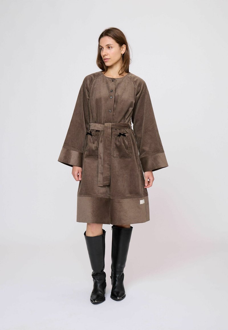 Nő barna kordbársony kabátot visel széles ujjakkal, derékszíjjal, elülső zsebekkel és fekete térdig érő csizmával, egyszínű háttér előtt áll.