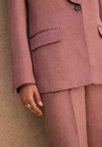 Ljus mauve skräddarsydd kostym med en knapp, två fickor och en slät textur. Har något utsvängda ärmar och inga synliga mönster.