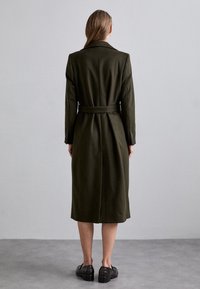 Cappotto in misto lana verde scuro con vita alta e maniche lunghe, con taglio dritto e spacco sul retro, abbinato a scarpe nere.