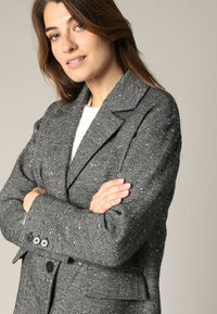 Grijze blazer met visgraatpatroon, voorzien van een notch-kraag, knoopmanchetten en twee voorzakken. Getailleerde pasvorm met gestructureerde stof.