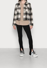 Karierte Jacke in Schwarz und Weiß, strukturierter beigefarbener Pullover, schwarze Hose mit seitlichen Schlitzen und weiße Sneaker mit schwarzen Akzenten.