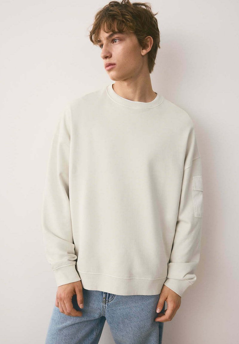 Jeune homme portant un sweat-shirt uni blanc cassé et un jean bleu clair, regardant sur le côté sur un fond clair uni.