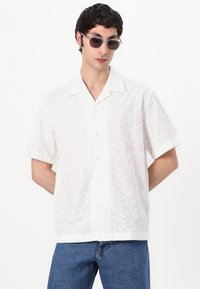 Camisa branca de mangas curtas com um padrão ondulado, feita de um tecido leve, com botões e um colarinho descontraído. Usada com jeans azuis.