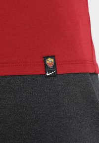 Camiseta de algodón roja con etiqueta negra de Nike y emblema de Roma, combinada con tela gris oscuro. Textura suave, diseño clásico y corte ajustado.