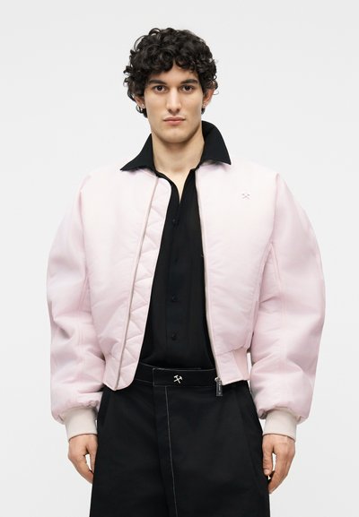 Jeune homme aux cheveux bouclés portant une doudoune rose clair par-dessus une chemise noire à boutons et un pantalon noir avec un petit logo.