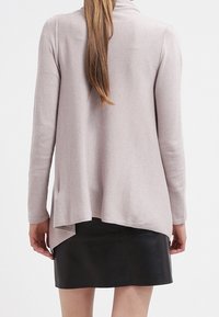 Lichtroze cardigan met een ontspannen pasvorm, open voorkant en een langere achterzoom, gecombineerd met een zwarte leren rok. Gladde textuur, minimale details.