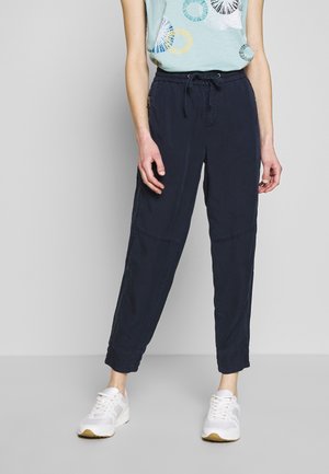 Persoon gekleed in zwarte broek met trekkoord, lichtblauw topje met patroon en witte sneakers, staand tegen een effen lichtgrijze achtergrond.