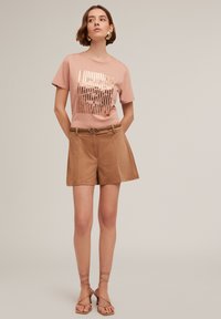 Tricou roz deschis cu un design grafic metalic, asortat cu pantaloni scurți bej cu talie înaltă și sandale bej cu barete. Textile cu texturi netede.