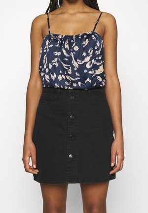 Femme portant un haut sans manches bleu marine à motifs et une jupe noire boutonnée, debout devant un fond blanc uni.