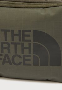 Τσάντα από ελαιόχρωμο ύφασμα με σχέδιο πλέγματος και μεγάλο μαύρο λογότυπο. Το υλικό είναι ανθεκτικό και σχεδιασμένο για εξωτερική χρήση.