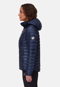 Kvinna i marinblå täckjacka med huva och logotyp på överarmen, stående på sidan mot en enfärgad vit bakgrund.