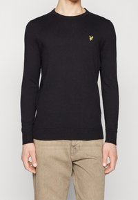 Pull en maille noire à manches longues, col rond, poignets et ourlet côtelés. Présente un petit logo aigle jaune sur la poitrine.