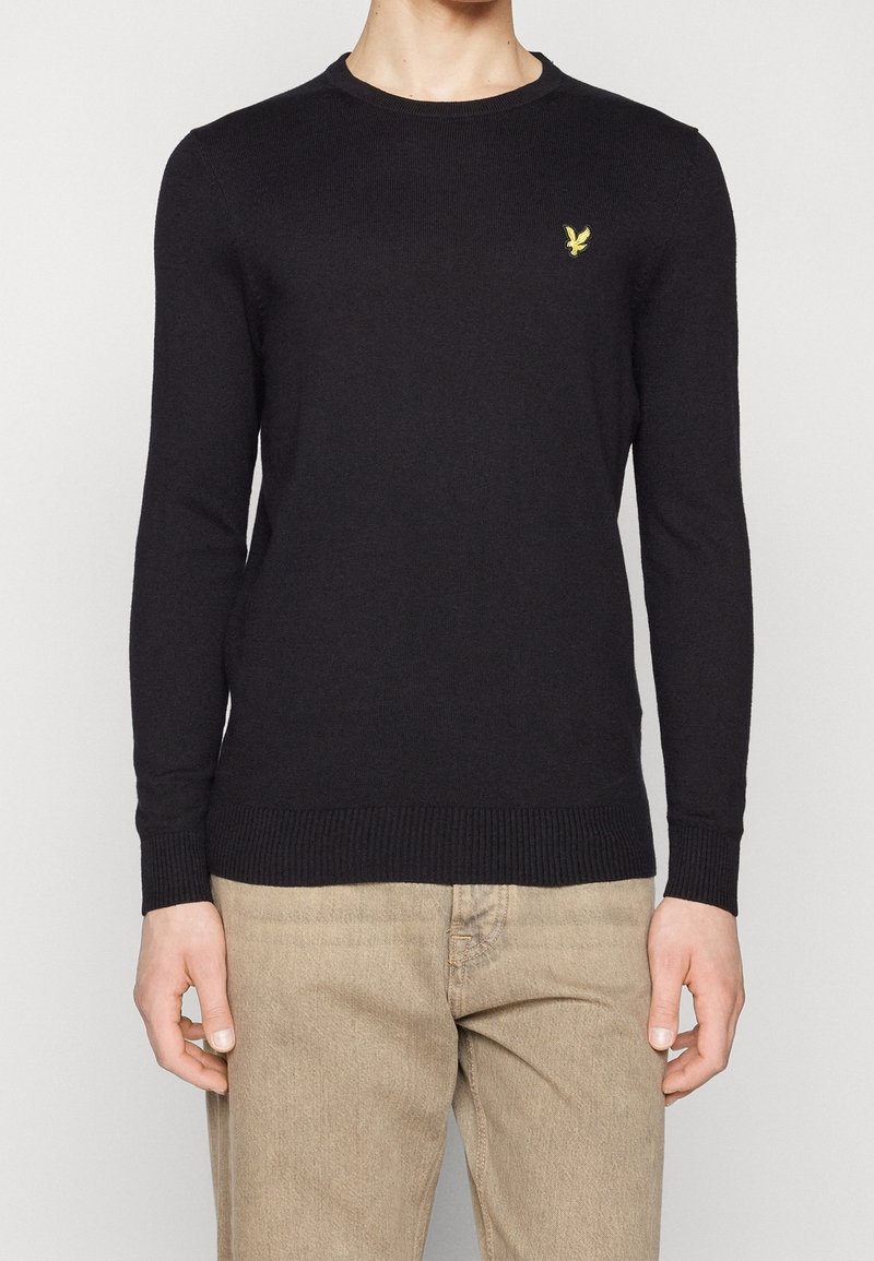 Pull en maille noire à manches longues, col rond, poignets et ourlet côtelés. Présente un petit logo aigle jaune sur la poitrine.