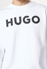 Sudadera blanca de algodón con un gran logo negro "HUGO" en el pecho. Diseño clásico de cuello redondo con mangas largas.