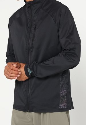 Homme portant une veste noire zippée avec une petite poche zippée sur la poitrine et le logo Adidas sur le poignet gauche.
