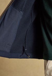 Innenansicht einer dunkelblauen Fleecejacke mit Kordelzug am Saum und teilweise sichtbarem grünem Ärmel und beiger Cordhose.