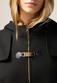 Abrigo negro con cremallera y capucha, que presenta una cremallera dorada y detalles de cuero. La textura parece suave con un acabado liso.