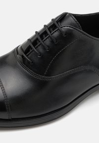 Zapato de cuero negro con punta redondeada, sutiles acentos de costura y cierre de cordones. Textura suave y diseño minimalista elegante.