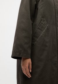 Right hand relaxed by side wearing dark olive long-sleeve coat with slanted side pocket on light background.