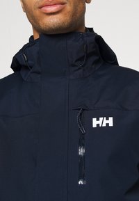 Chaqueta impermeable de color azul marino con un alto cuello. Cuenta con un bolsillo frontal con cremallera, un cordón ajustable y un logo blanco en el pecho. Textura suave.
