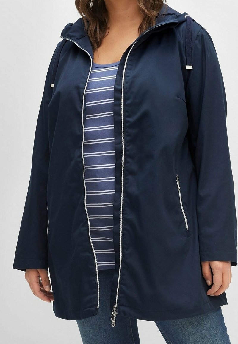 Marineblauw waterdicht jack met capuchon, ritssluiting aan de voorkant, zijritszakken en verstelbare koordjes. Gedragen over een gestreept shirt.