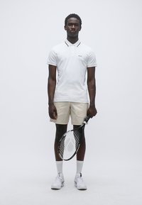 Λευκό πόλο μπλουζάκι με μαύρο περίγραμμα, μπεζ σορτς, λευκές κάλτσες και αθλητικά παπούτσια. Κρατάει ρακέτα τένις. Απλός σχεδιασμός, χωρίς σχέδια.