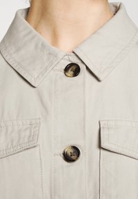 Beżowa kurtka z kołnierzem z dwoma przednimi kieszeniami, z czarnym guzikiem o marmurkowym wzorze, wykonana z fakturowanej tkaniny.