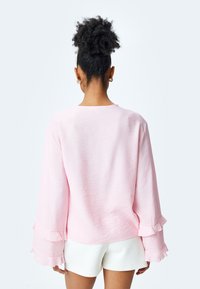 Maglietta rosa a maniche lunghe con polsini arricciati, tessuto leggero, scollatura tonda e vestibilità comoda, abbinata a pantaloni corti bianchi.
