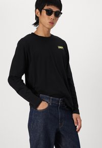 Long-sleeve black T-shirt with a yellow “CNCT” logo, paired with dark blue denim jeans. Person wearing black sunglasses. Plain background.
