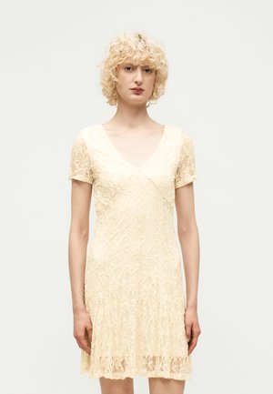 Femme aux cheveux blonds bouclés portant une robe courte en dentelle crème avec un col en V, debout devant un fond clair uni.