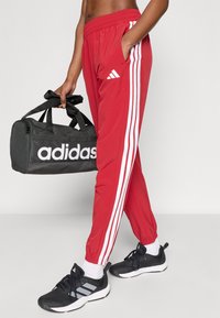 Κόκκινο αθλητικό παντελόνι με λευκές ρίγες και ελαστική μέση, σε συνδυασμό με μαύρη τσάντα duffel adidas και μαύρα αθλητικά παπούτσια.