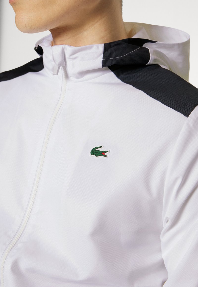 Veste légère blanche avec des panneaux d'épaule noirs, fermeture éclair à l'avant, et un logo de crocodile vert sur la poitrine gauche. Tissu lisse et texturé.