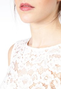 Biała koronkowa bluzka z misternymi wzorami kwiatowymi i przezroczystą strukturą, z wysokim dekoltem i bezrękawowym fasonem.