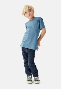 Sininen puuvillainen t-paita lyhyillä hihoilla, yhdistetty tummansinisiin farkkuihin ja vihreisiin lenkkareihin. Yksinkertainen malli ilman kuvioita tai merkittäviä koristeita.