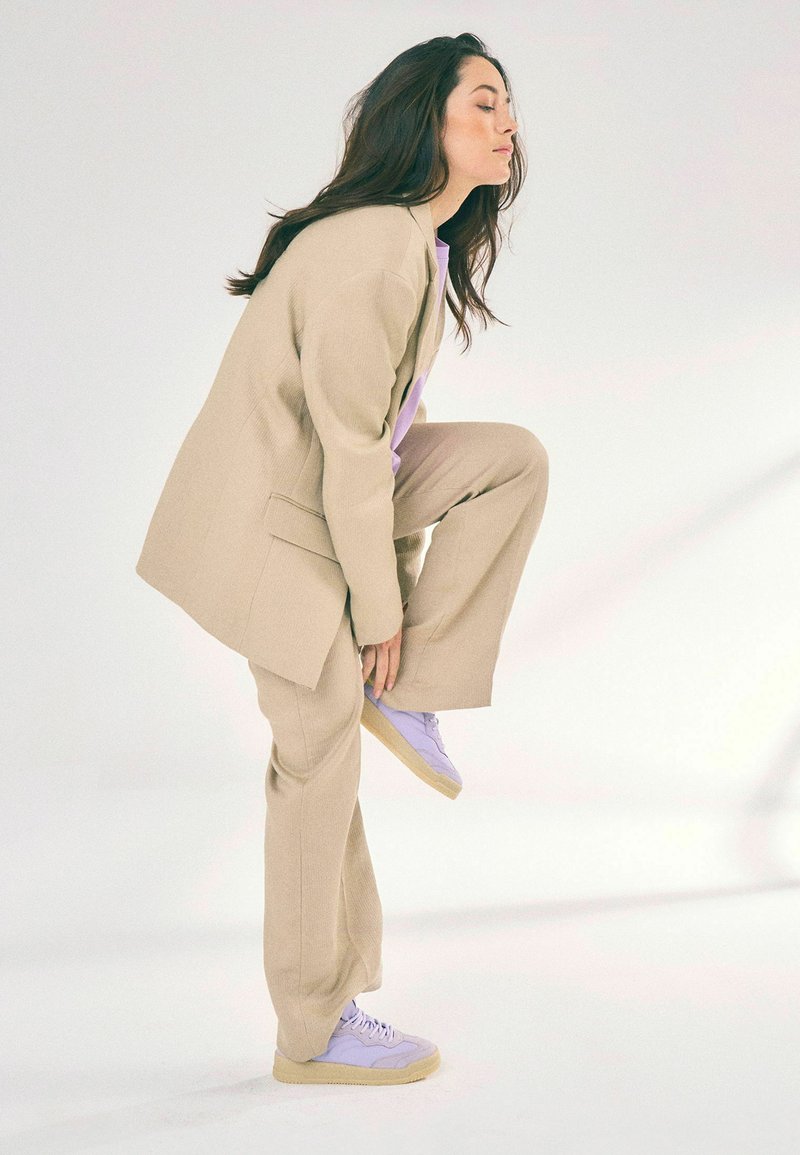 Beige tailored suit with a textured finish, fitted pants, and a lilac top. Purple sneakers with a light sole accentuate the outfit.