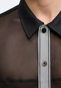 Μαύρο δίχτυ πουκάμισο με απλό μαύρο γιακά και λευκή λωρίδα κουμπιών με δύο μαύρα κουμπιά, φορεμένο σε γυμνό δέρμα.