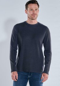 Dunkelgraues Langarmshirt aus strukturiertem Stoff mit Rundhalsausschnitt und dezenten Schulterdetails, kombiniert mit blauen Jeans.