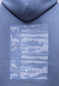 Μπλε φούτερ με κουκούλα, με ένα μεγάλο ορθογώνιο μπαλώμα στην πλάτη, που απεικονίζει ένα αφηρημένο εικονοστοιχειωτό σχέδιο σε πιο ανοιχτές μπλε αποχρώσεις.