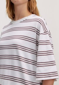 Weißes, oversized T-Shirt mit horizontalen bordeauxfarbenen Streifen, rundem Ausschnitt und kurzen Ärmeln. Das Material wirkt weich mit einer glatten Textur.