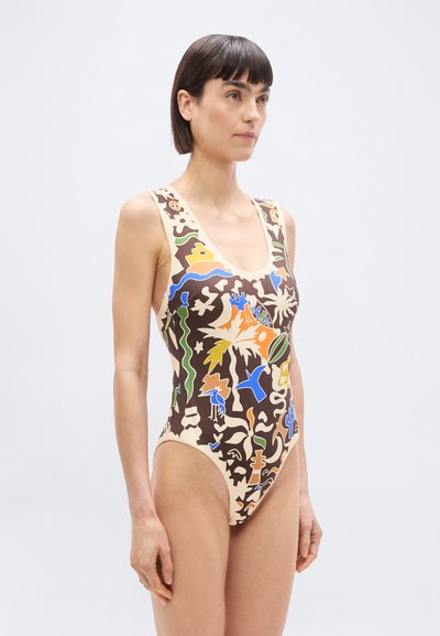 Γυναίκα που φοράει ολόσωμο μαγιό με πολύχρωμα αφηρημένα σχήματα και τροπικά μοτίβα σε καφέ και μπεζ φόντο, στέκεται μπ ροστά σε λευκό φόντο.