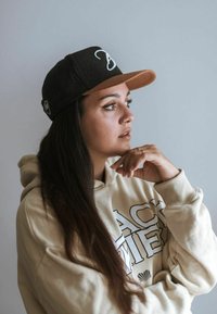 Cappello nero e marrone con logo, felpa beige con testo stampato. Lunghi capelli scuri e una mano appoggiata sul mento, sfondo chiaro.